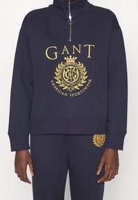 Sudadera de la marina de guerra con cuello alto, cremallera de cuarto y logo dorado que presenta un diseño de laurel. Puños y dobladillo acanalados, material de algodón suave.
