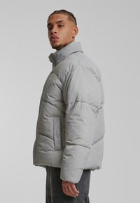 Chaqueta acolchada gris claro con un cuello alto, textura acolchada y bolsillos laterales. Presenta un diseño elegante y un ajuste ligeramente holgado.