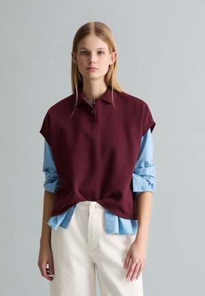 Młoda kobieta o prostych blond włosach, ubrana w bezrękawnik bordowy polo nałożony na niebieską koszulę w paski oraz białe spodnie, stoi na tle jednolitego szarego tła.