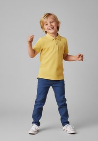 Uśmiechnięty młody chłopiec z blond włosami, ubrany w żółtą koszulkę polo, niebieskie spodnie i białe trampki, stojący z uniesionymi rękami.