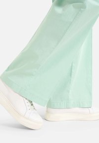 Baskets compensées blanches portées avec un pantalon ample vert clair, montrant un mouvement de marche sur un fond blanc.