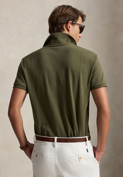 Mann iført solbriller, olivengrønn poloskjorte med oppslag på kragen, hvite bukser og brunt belte, vendt bort med hendene i lommene.