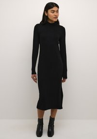 Schwarzes, geripptes Strickkleid mit hohem Kragen, langen Ärmeln und seitlichen Schlitzen, kombiniert mit knöchelhohen schwarzen Absatzstiefeln.