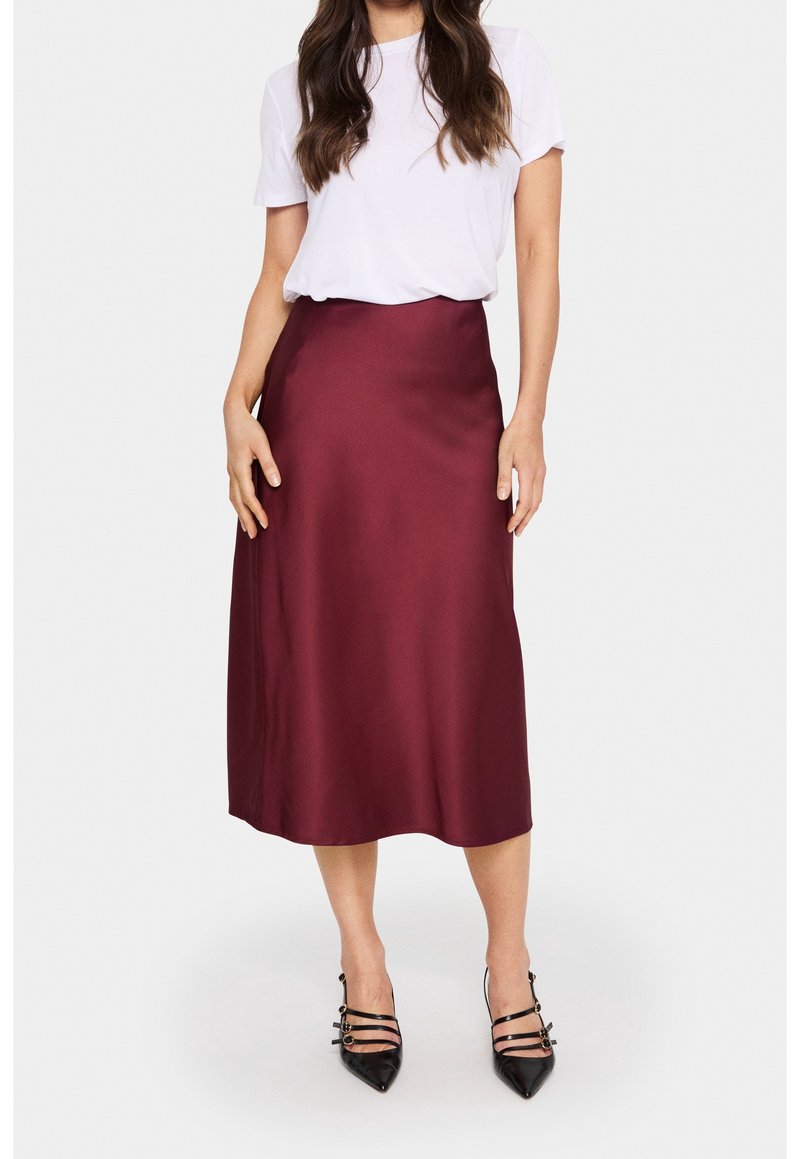Femme portant un haut blanc à manches courtes, une jupe midi bordeaux et des talons noirs pointus avec brides, debout devant un fond clair uni.