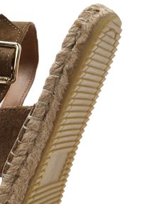 Sandalo in suede marrone con suola avvolta in juta. Presenta un tacco testurizzato, dettagli intrecciati e una fibbia regolabile liscia.