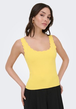 Femme portant un haut jaune côtelé sans manches à bords festonnés et un pantalon noir, posant avec une main sur la hanche sur un fond clair.