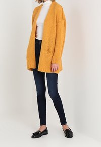 Mustard yellow knitted cardigan with open front, textured fabric, two front pockets, paired with dark skinny jeans and black loafers.