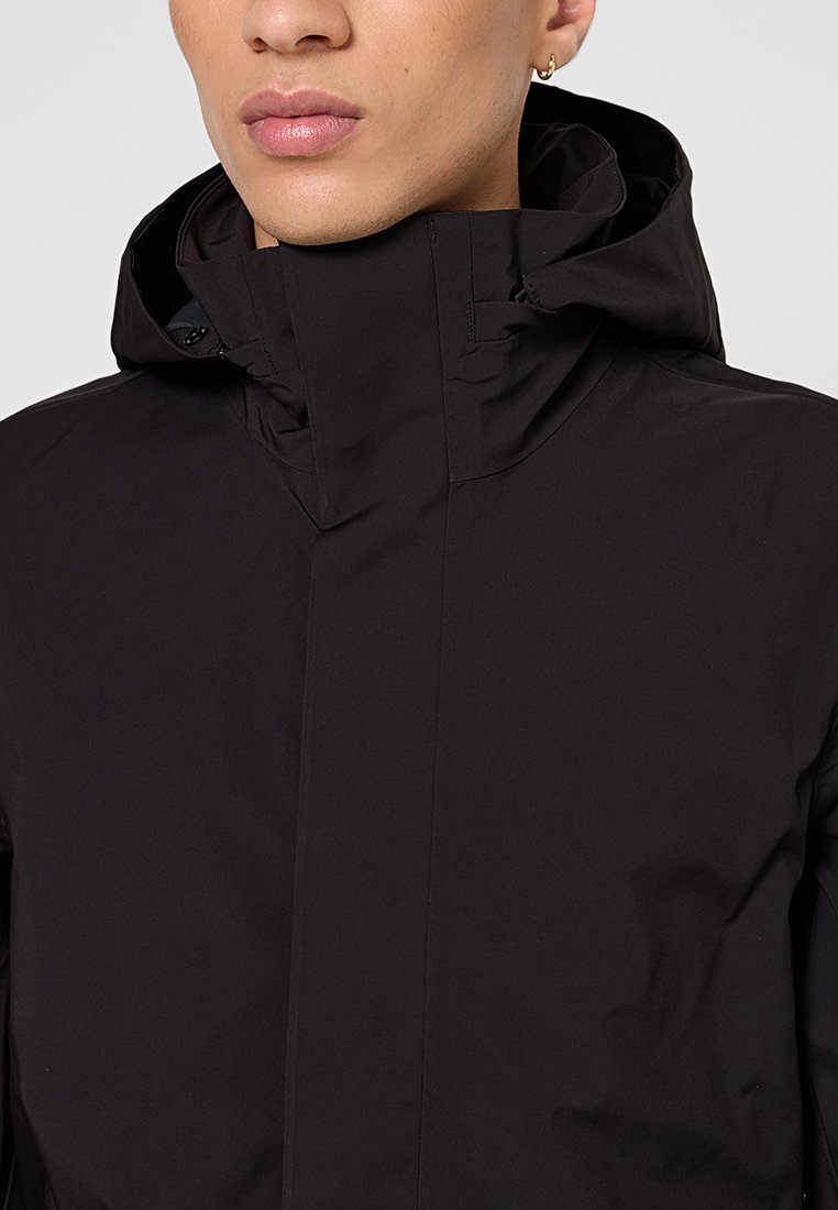 Junge Person mit schwarzer Kapuzenjacke mit hohem Kragen, die Lippen, Kinn und einen kleinen Ohrring in der linken Ohr zeigt.