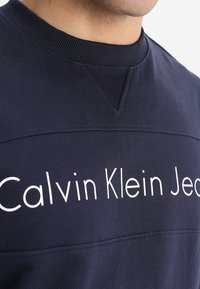 Felpa girocollo blu navy con scritta bianca "Calvin Klein Jeans", tessuto testurizzato e collo a coste con dettagli di cuciture evidenti.