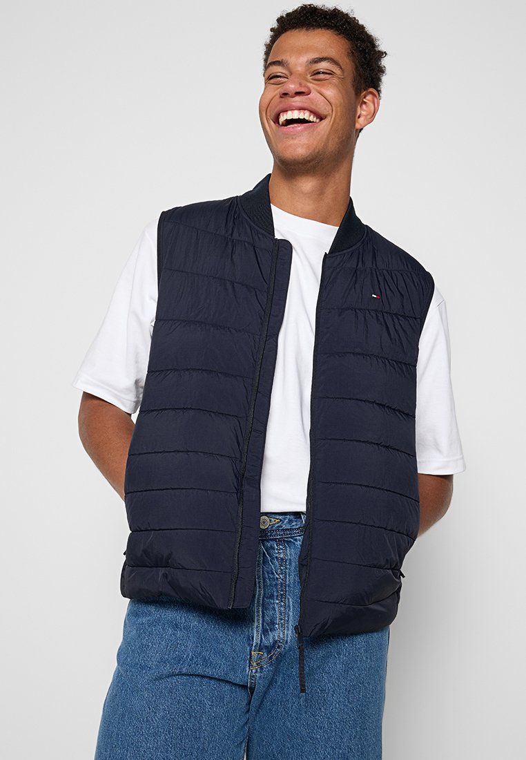 Chaleco acolchado azul marino con acolchado horizontal, cuello redondo acanalado y cremallera frontal. Lleva puesto un t-shirt blanco y está combinado con jeans de mezclilla azul.