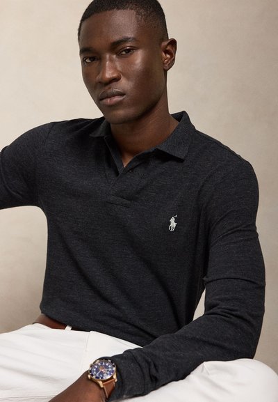 Vyras dėvi tamsų ilgomis rankovėmis polo marškinėlį su baltu siuvinėtu logotipu ir laikrodį su ruda odine apyranke, sėdintis prieš neutralią fono spalvą.