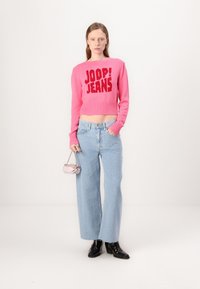 Svijetloružičasti pleteni skraćeni pulover sa natpisom "JOOP! JEANS" u podebljanim slovima, uparen s svijetloplavim širokim trapericama i malom metalik ružičastom torbicom.