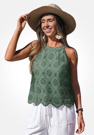 Vrouw glimlachend, met een hoed met brede rand, groene geborduurde mouwloze top, witte broek en kwastjesoorbellen, die haar hoed met één hand aanpast.