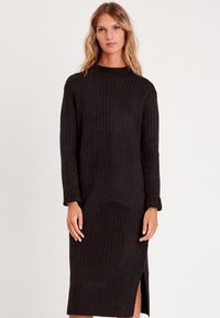 Vestido negro de punto acanalado con mangas largas, cuello alto y abertura lateral. Presenta una tela texturizada con un corte slim y recto.