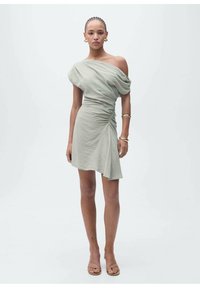 Off-shoulder, light green dress with draped fabric, side ruching, asymmetrical hem, and smooth texture. Worn with simple sandals and accessories.