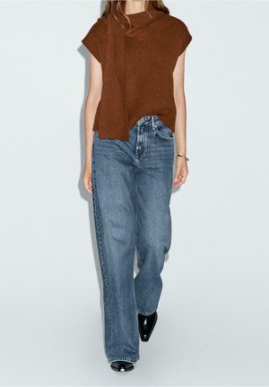 Femme portant un haut en maille marron sans manches, un jean bleu large, des chaussures noires à bouts pointus et un bracelet en or au poignet droit.