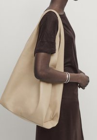 Personne portant un haut marron foncé manches courtes et une jupe, portant un grand sac cabas beige sur l'épaule, avec des bracelets argentés.