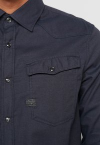 Mörk marinblå skjorta med knappar gjord av strukturerad tyg, med spetsig krage, en bröstficka och ett märke på framsidan.