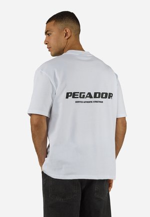 Mann trägt ein weißes kurzärmeliges T-Shirt mit dem Text "PEGADOR Certified Authentic Streetwear" auf der Rückseite, kombiniert mit schwarzen Hosen, Rücken zum Betrachter gewandt.