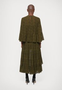 Osoba s oholeními vlasy, oblečená v dlouhé, olivově zelené šaty s paisley vzorem a rozšířenými rukávy, a černými podpatky, stojící zády k fotoaparátu.
