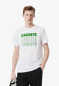 Joven vestido con una camiseta blanca con el texto verde "LACOSTE", sosteniendo una prenda a juego en verde y blanco, de pie frente a un fondo liso.