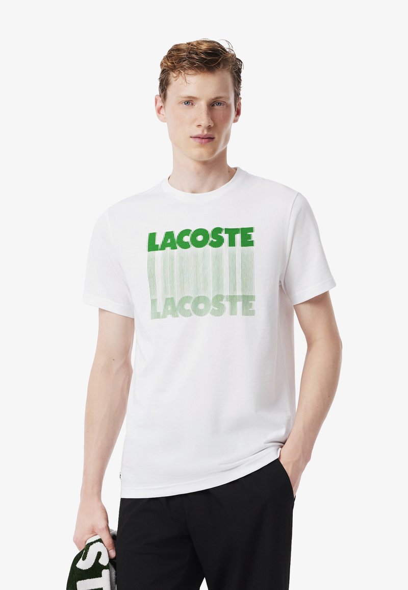 Joven vestido con una camiseta blanca con el texto verde "LACOSTE", sosteniendo una prenda a juego en verde y blanco, de pie frente a un fondo liso.