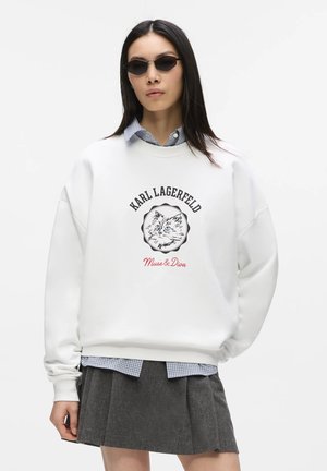 Kvinde iført mørke solbriller, hvid oversized sweatshirt med kattegrafik og "Karl Lagerfeld Muse & Diva" tekst, ternet skjorte og grå plisseret nederdel.