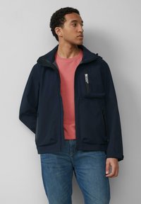 Granatowa wodoodporna kurtka z kieszenią na zamek błyskawiczny na klatce piersiowej i dwoma kieszeniami po bokach. Nosi się ją na różowej koszuli, w połączeniu z niebieskimi dżinsami.