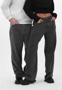 To personer har løstsiddende, mørkegrå denimbukser med forlommer. Den ene bærer en sort cropped top, mens den anden har en lys grå sweatshirt.