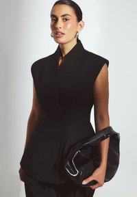 Zwarte getailleerde mouwloze top met een hoge kraag. Gecombineerd met een stijlvolle zwarte leren tas, die de nadruk legt op strakke lijnen en een gestructureerd ontwerp.