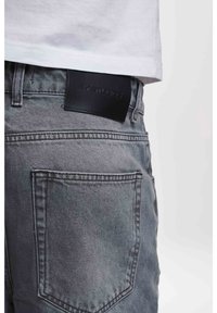 Šedé džíny z denimu s výraznou zadní kapsou, na černé kožené štítku na pasu s vypáleným textem, představující čistý, texturovaný povrch.