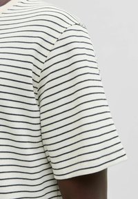 Hvid skjorte med korte ærmer og tynde sorte vandrette striber, båret af en person med mørk hud, vist fra skulder til midt på armen.