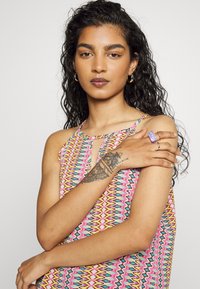 Top sem mangas multicolorido com um padrão em zigue-zague em rosa, azul e amarelo. Detalhe de decote em forma de olho-chave. Braços do modelo adornados com anéis e uma tatuagem.