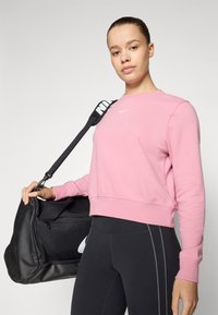 Sweatshirt rose avec des poignets côtelés, arborant un petit logo blanc. Sac de sport noir avec une sangle contrastante, tenu par une personne en leggings noirs.