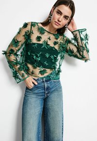 Grünes, durchsichtiges Oberteil mit bestickten floralen Details und langen Ärmeln, kombiniert mit hellblauen weiten Jeans. Strukturiertes Design mit schichtartigen Elementen.