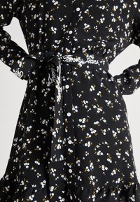 Černé košilové šaty s bílými a modrými květinovými vzory. Mají zavazovací pas s logem "Tommy Jeans" a volánkový lem.