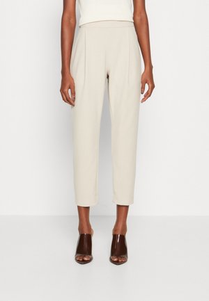 Pantalons beiges taille haute avec plis, longueur cheville et poches latérales. Tissu lisse avec une coupe structurée. Portés avec des talons ouverts bruns.