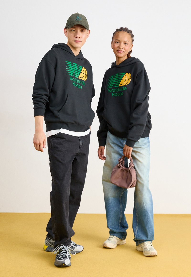 Czarne bluzy z kapturem z zielonymi i żółtymi grafikami, czarne dżinsy, niebieskie dżinsy z jasnym przetarciem, szara czapka i mała fioletowa torba.