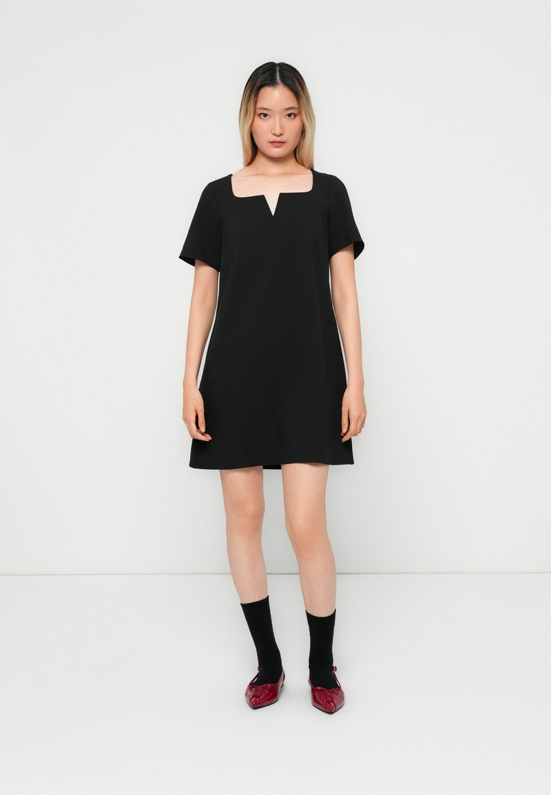 Black short-sleeve dress with a square neckline and v-shaped cutout. Smooth fabric, knee-length, paired with black socks and red shoes.