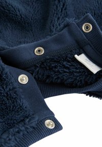 Marinblå fleece tyg med en mjuk, strukturerad yta. Har silverfärgade tryckknappar och en tygetikett som delvis är synlig vid kanten.