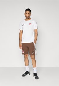 Hvid kortærmet t-shirt med logo, parret med brune shorts med sidelogo og striber. Sorte træningssko og hvide sokker.