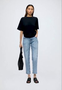 Žena nosící černé oversized tričko, světle modré rovné džíny, černé mule boty s otevřenou špičkou a držící černou kabelku na jednobarevném pozadí.