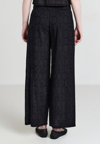 Schwarzweite Hose mit kreisförmigen gestickten Mustern, getragen mit passenden strukturierten schwarzen Flats, von hinten auf einem hellen Hintergrund gezeigt.