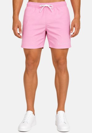 Homme portant un short de bain rose clair avec cordon blanc et baskets blanches, debout devant un fond uni.