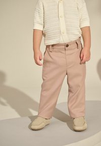 Hellrosa Hose mit einem Frontknopf und Gürtelschlaufen, kombiniert mit einem cremefarbenen kurzärmligen Strickcardigan über einem weißen Hemd; beigefarbene Schuhe.