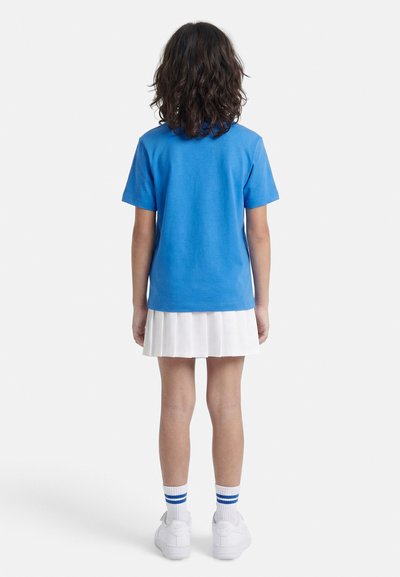 Παιδί με σγουρά μαλλιά μέχρι τον ώμο, φοράει μπλε κοντομάνικο πουκάμισο, λευκή πλισέ φούστα, λευκές κάλτσες με μπλε ρίγες και λευκά αθλητικά παπούτσια.