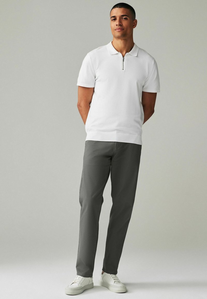 Polo bianco con colletto a zip corto, abbinato a pantaloni grigi e sneakers bianche. Tessuto dalla texture liscia, design su misura.