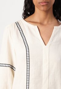 Off-white bluze no teksturēta auduma ar V veida kakla izgriezumu, garām piedurknēm un melnu ģeometrisku izšūtu rakstu pa centru un piedurknēm.