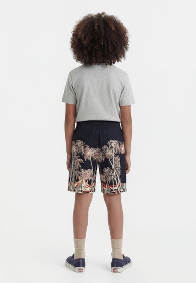 Enfant debout, dos tourné, portant un t-shirt gris, un short bleu marine avec un imprimé de palmiers, des chaussettes beiges et des baskets bleu marine.