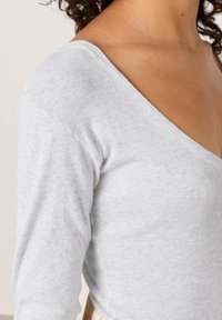 Svijetlosivi rebrasti top s dugim rukavima koji ima duboki V-izrez i uski kroj, prikazujući mekani, teksturirani materijal i suptilne šavove.